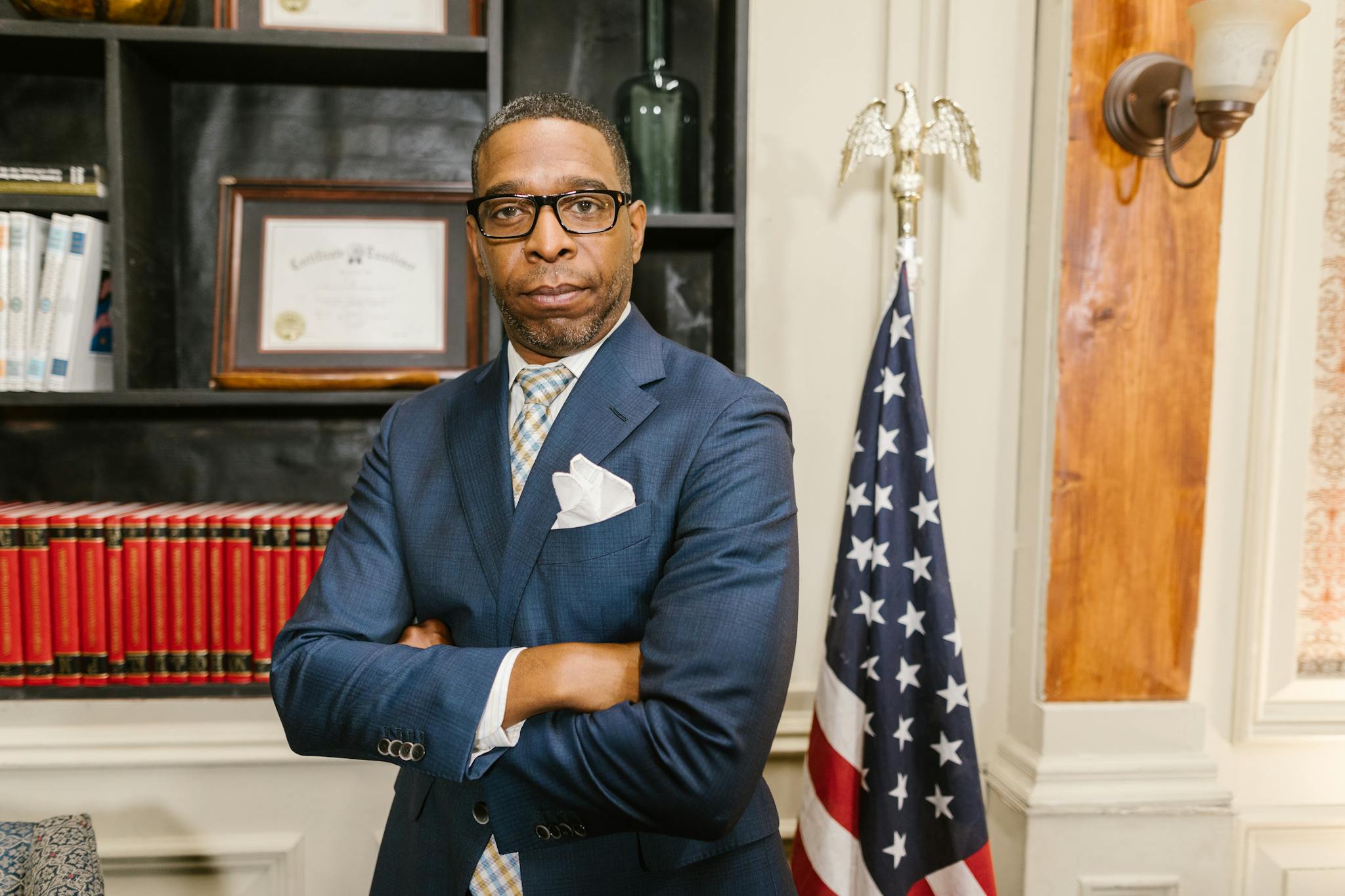 Portrait of a confident lawyer in office attire, standing with arms crossed in front of a USA flag.